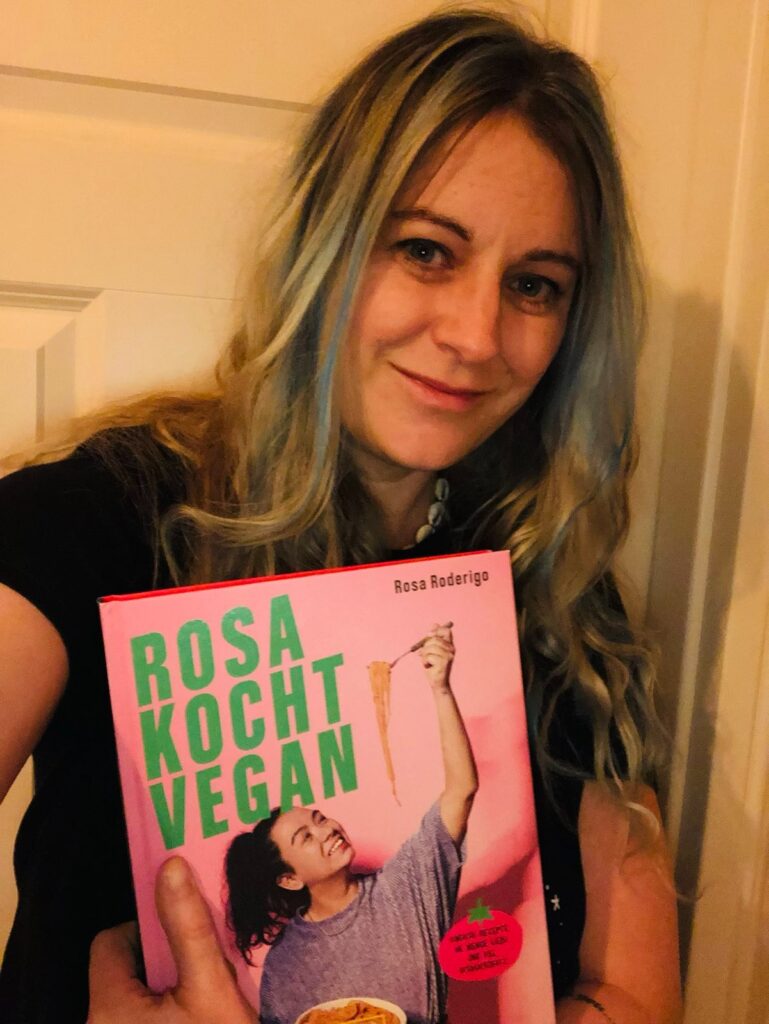 Foto: Eine weiße Frau mit blauen Strähnchen in den Haaren hält das Kochbuch "Rosa kocht vegan" hoch. Auf dem rosa Cover zieht eine asiatische Frau mit nach oben ausgestrecktem Arm Spaghetti lang.